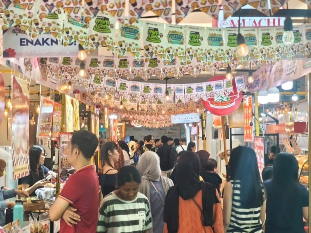 Suasana keramaian pengunjung di lorong tenant makanan dan fashion MTF Market Fasting Forward di Mall Phinisi Point Makassar, dihiasi dekorasi lampu gantung dan bendera kecil warna-warni.
